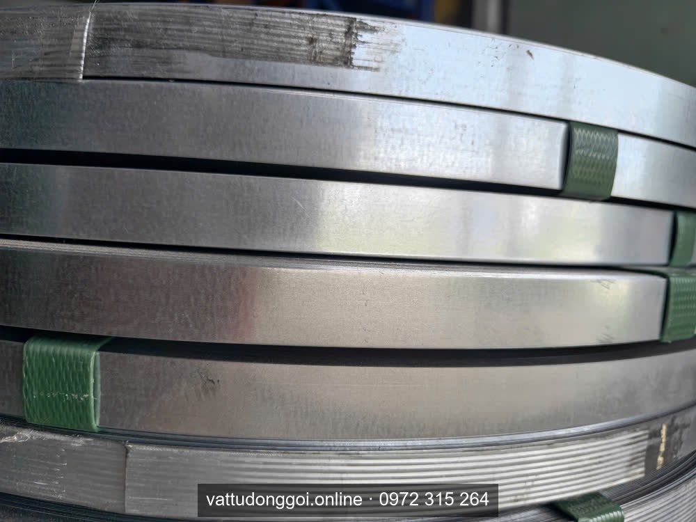 Cạnh các cuộn đai thép mạ kẽm xếp chồng, có miếng nhựa xanh kẹp giữa các cuộn