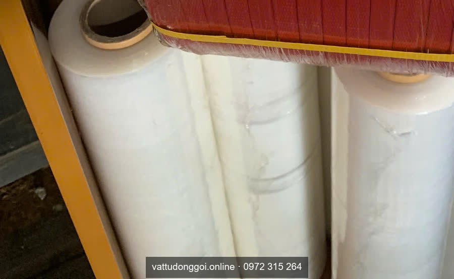 Cuộn màng PE quấn pallet màu trắng đặt trên kệ, bên dưới cuộn dây đai đỏ